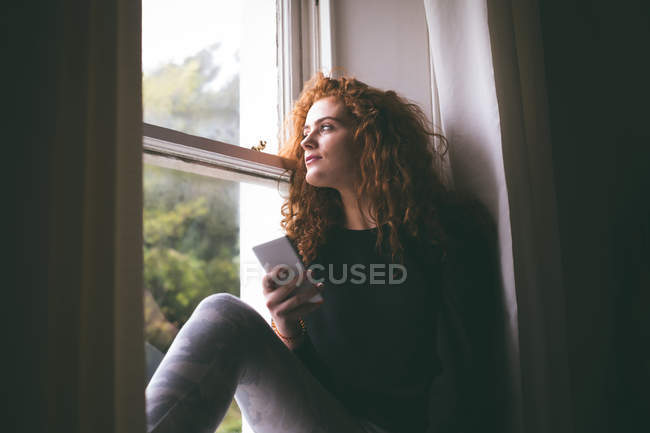 Mujer reflexiva con teléfono móvil sentado en el alféizar de la ventana en casa - foto de stock