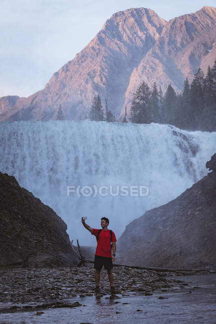 Young man taking selfie with mobile phone near waterfall — Stock Photo