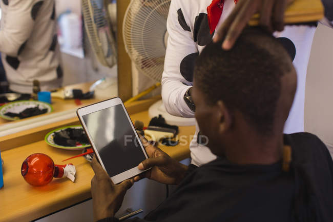 Customer using digital tablet while barber trimming his hair in barber shop — Stock Photo