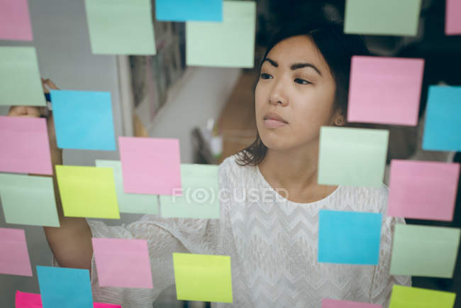 Exécutif féminin collant des notes adhésives sur le mur de verre dans le bureau — Photo de stock