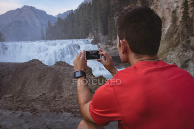 Rear view of man taking photo of waterfall with mobile phone — Stock Photo