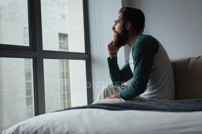 Hombre mirando a través de la ventana en el dormitorio en casa - foto de stock