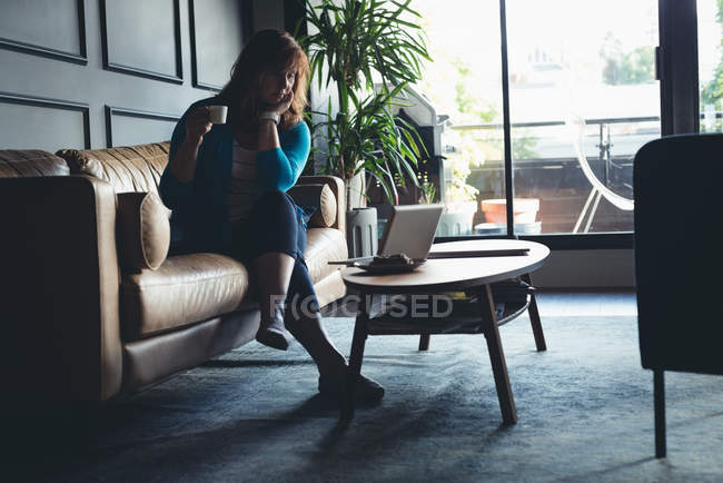 Empresária tomando café enquanto usa laptop no escritório — Fotografia de Stock
