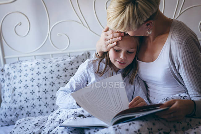 Любящая мать целует свою дочь во время чтения книги в спальне — стоковое фото