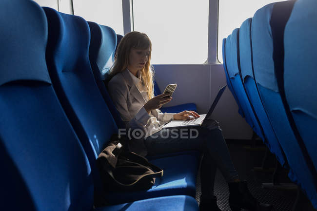 Жінка перевіряє мобільний телефон під час використання ноутбука на круїзному кораблі — стокове фото