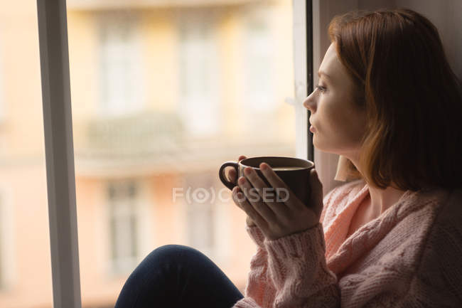 Femme prenant un café tout en regardant par la fenêtre à la maison — Photo de stock