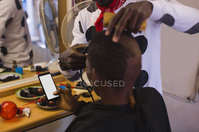 Client utilisant un téléphone portable tout barbier couper ses cheveux dans le salon de coiffure — Photo de stock