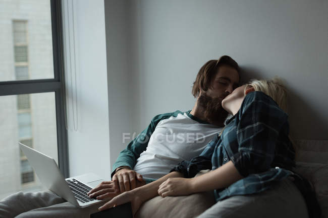 Casal romântico beijando uns aos outros ao usar laptop em casa — Fotografia de Stock
