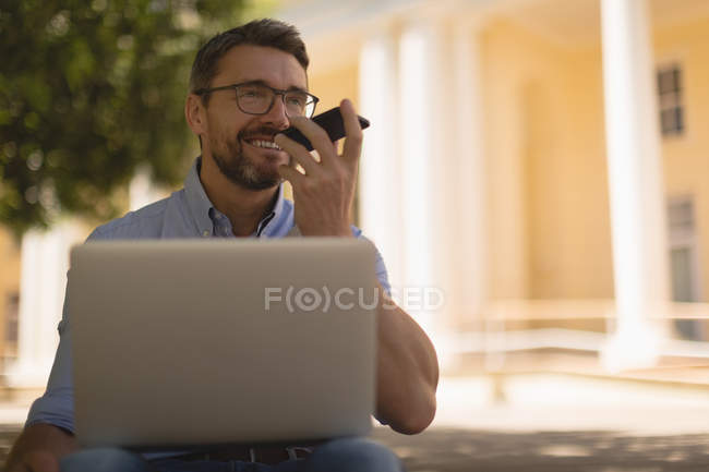 Homme parlant sur un téléphone mobile tout en utilisant un ordinateur portable dans le parc — Photo de stock