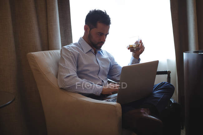 Homme d'affaires utilisant un ordinateur portable tout en ayant whisky dans la chambre d'hôtel — Photo de stock