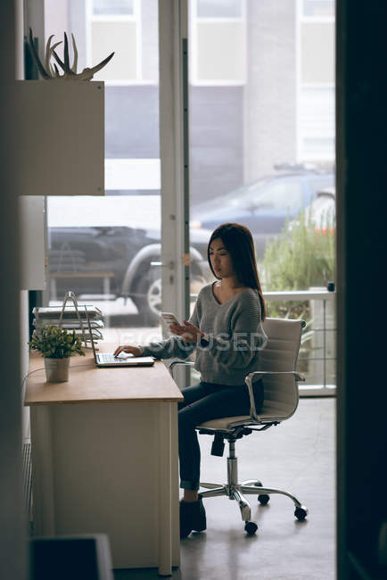 Жіночий виконавчий використання мобільного телефону під час роботи над ноутбук на столі в офісі — стокове фото