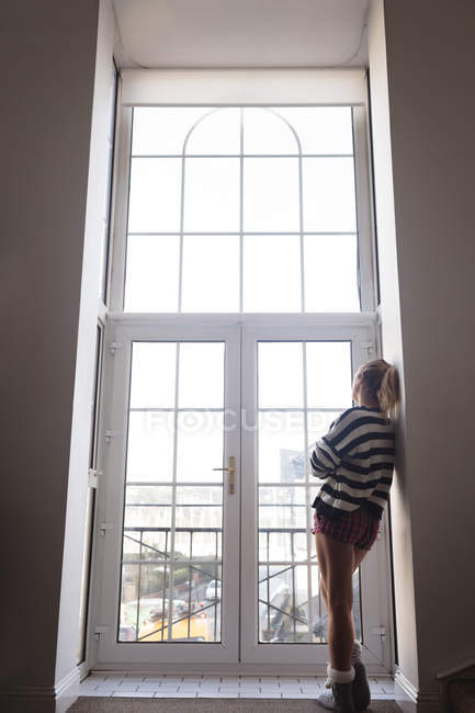Vista trasera de la mujer reflexiva mirando a través de la ventana en casa - foto de stock