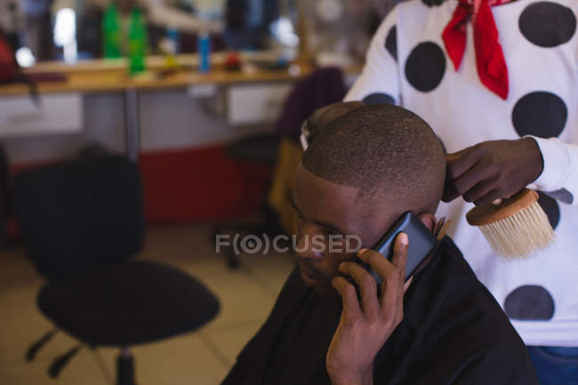 Клієнт розмовляє на мобільному телефоні, коли перукар обрізає волосся в перукарні — стокове фото