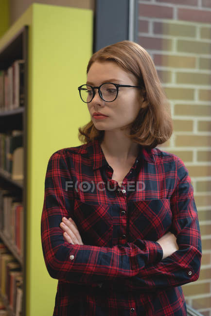 Задумчивая женщина, стоящая со скрещенными руками в библиотеке — стоковое фото
