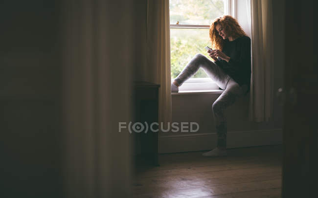 Женщина, использующая мобильный телефон, сидя дома на подоконнике — стоковое фото