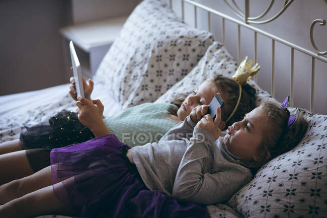Братья и сестры с помощью мобильного телефона и цифрового планшета на кровати в спальне — стоковое фото