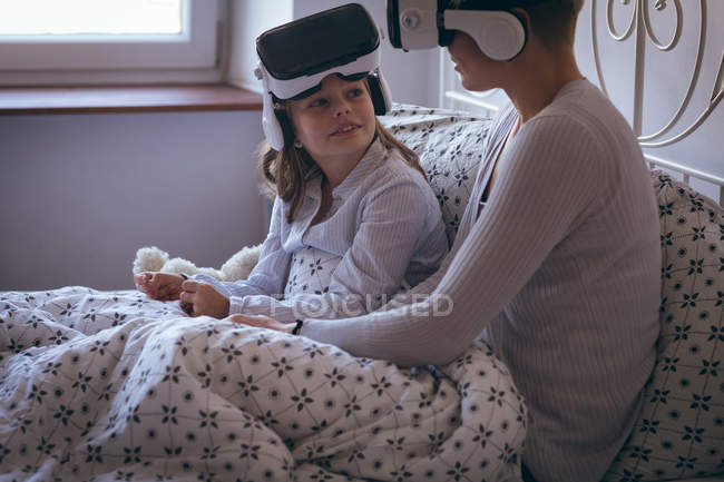 Madre e hija interactuando mientras usan auriculares de realidad virtual en la cama - foto de stock