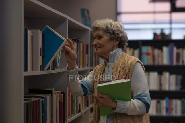 Mujer mayor quitando libro de estante de libro en la biblioteca - foto de stock