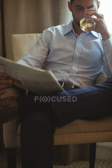 Empresário lendo jornal enquanto bebe uísque no quarto de hotel — Fotografia de Stock