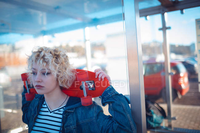 Mujer reflexiva sosteniendo monopatín en la parada de autobús - foto de stock