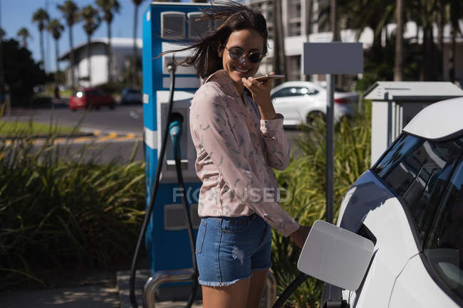 Mulher falando no telefone celular enquanto carrega carro elétrico na estação de carregamento — Fotografia de Stock