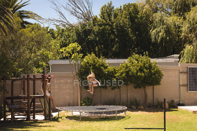 Девушка прыгает на батуте в саду в солнечный день, сестра смотрит — стоковое фото