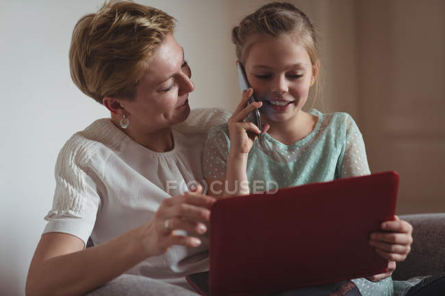 Мати і дочка розмовляють на мобільному телефоні, використовуючи ноутбук у вітальні вдома — стокове фото