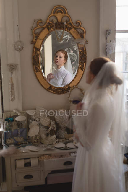 Біла наречена у весільній сукні та вуалі, дивлячись у дзеркало на старовинному бутіку — стокове фото