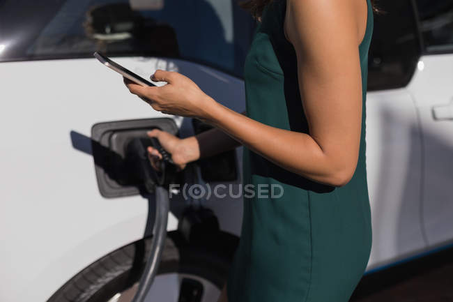 Sección media de la mujer que usa el teléfono móvil mientras carga el coche eléctrico en la estación de carga - foto de stock
