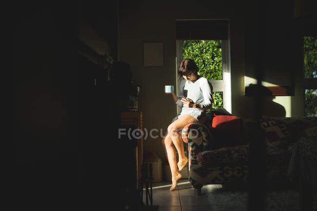 Mulher tomando café ao usar o telefone celular na sala de estar em casa — Fotografia de Stock