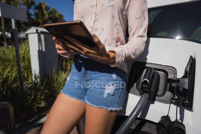 Sección media de la mujer que usa la tableta digital mientras carga el coche eléctrico en la estación de carga - foto de stock