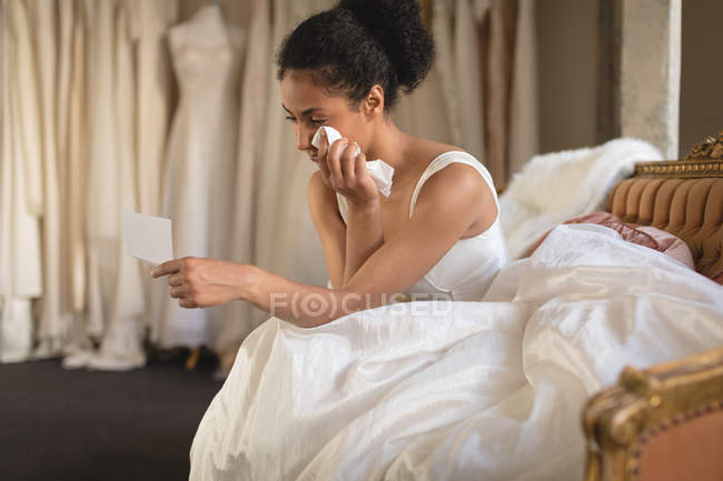 Jeune mariée essuyant des larmes avec du papier mouchoir assis sur le canapé et lisant la carte de vœux — Photo de stock