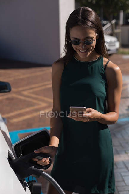 Mujer usando el teléfono móvil mientras carga el coche eléctrico en la estación de carga - foto de stock