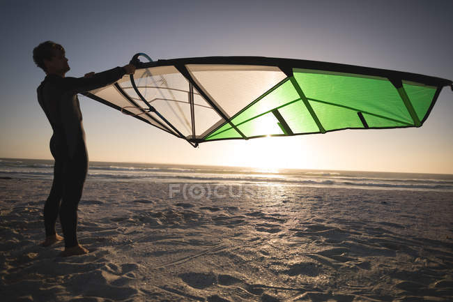 Surfista masculino segurando um papagaio na praia ao entardecer — Fotografia de Stock