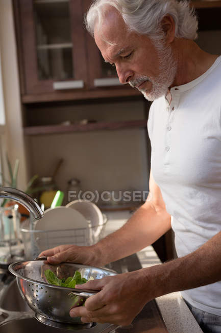Hombre mayor limpieza vegetal con agua en la cocina en casa - foto de stock