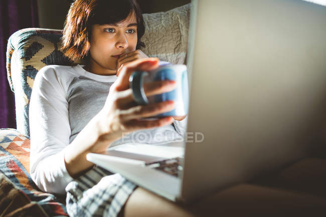 Mujer tomando café mientras usa el ordenador portátil en la sala de estar en casa - foto de stock