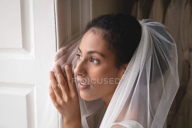 Retrato de noiva em vestido de noiva e véu olhando através da janela — Fotografia de Stock
