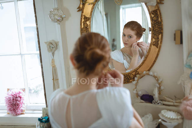 Молода наречена у весільній сукні носить сережки на бутіку і дивиться у дзеркало на стіну — стокове фото