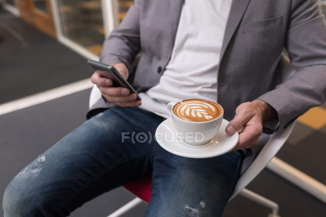 Mid section of businessman having coffee while using mobile phone in offic — Stock Photo