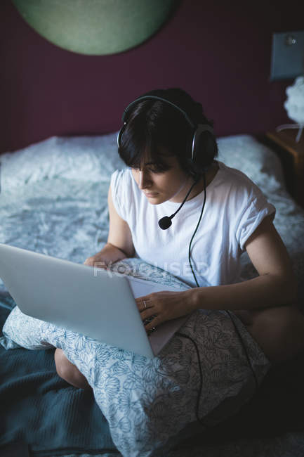 Femme utilisant un ordinateur portable avec casque dans la chambre à coucher à la maison — Photo de stock
