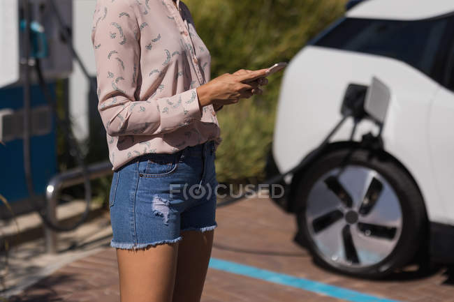 Sección media de la mujer que usa el teléfono móvil mientras carga el coche eléctrico en la estación de servicio - foto de stock