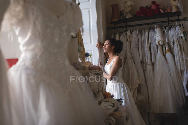 Mujer de raza mixta, novia en vestido blanco mirando a través de la ventana en la boutique - foto de stock
