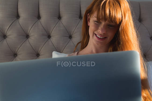 Улыбающаяся женщина с рыжими волосами с помощью ноутбука в спальне дома — стоковое фото