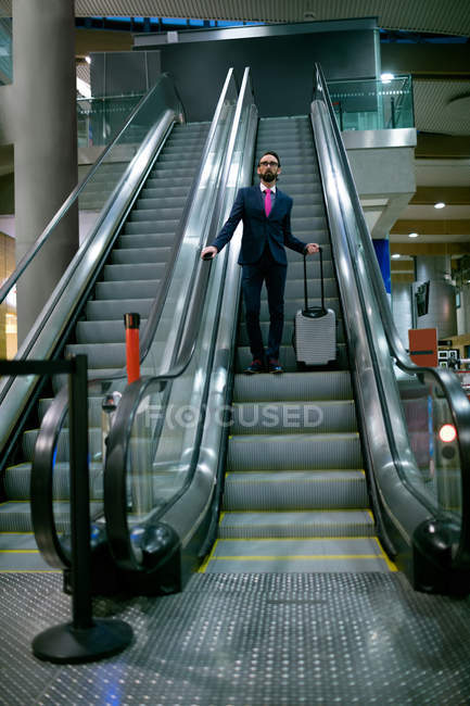 Бизнесмен, стоящий на эскалаторе с багажом в аэропорту — стоковое фото