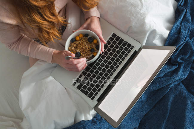 Donna con capelli rossi utilizzando il computer portatile in camera da letto con colazione ciotola — Foto stock