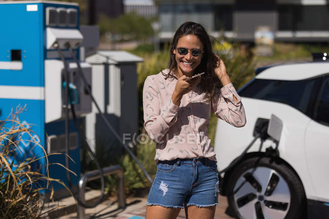 Женщина разговаривает по мобильному телефону во время зарядки электромобиля на зарядной станции — стоковое фото