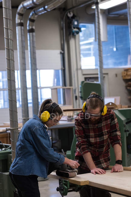 Женщина плотник с помощью самолета Джек в то время как мужчина смотрит на нее в мастерской — стоковое фото
