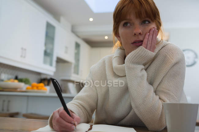 Thoughtful woman writing on diary at home in kitchen with hand on cheek — Stock Photo