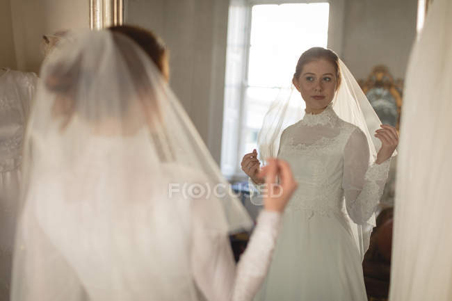 Novia caucásica en vestido de novia y velo mirando en el espejo en boutique vintage - foto de stock