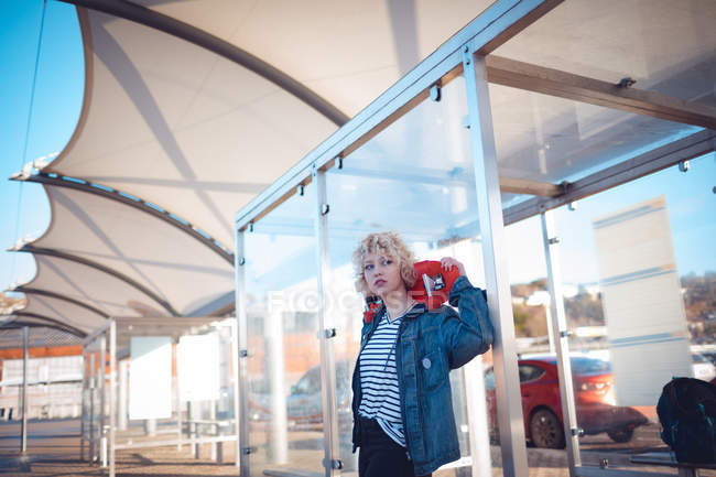 Задумчивая женщина держит скейтборд на автобусной остановке — стоковое фото